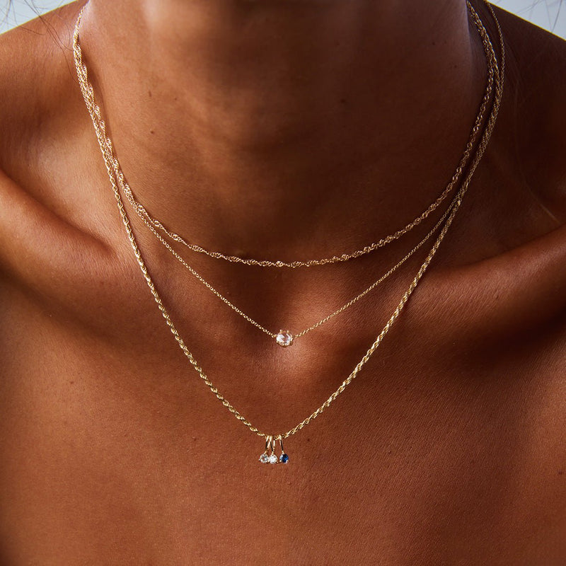 A close-up shot features a person adorned with three gold necklaces of varying lengths. The shortest necklace is an elegant gold chain, the middle one is highlighted by a small round pendant, and the longest showcases three delicate tear-drop shaped charms. The background is softly blurred in neutral tones. All necklaces are part of the "supernova necklace" collection by bluboho, crafted in 14k yellow gold with white diamonds.