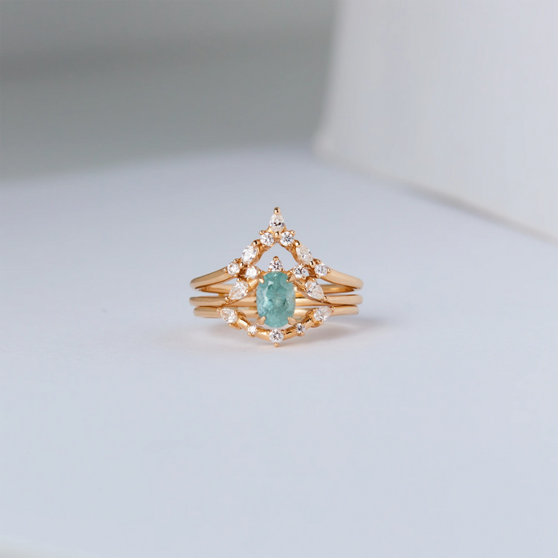 A close-up of the Love's Unity One-of-a-Kind ring set by Beloved by Bluboho, featuring a cyan green oval sapphire center stone surrounded by intricate diamond accents. The 14k yellow gold rings are stacked together, forming a delicate and elegant design, placed on a white background.