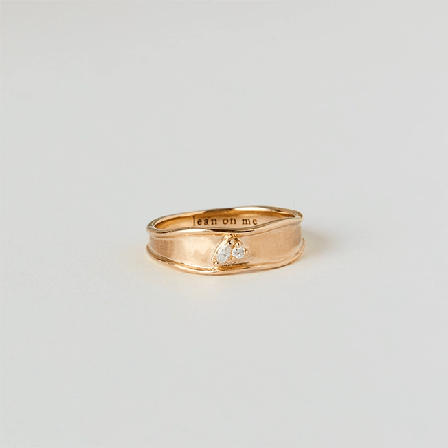 Introducing the kindred spirits lean on me band ring from bluboho, crafted in 14k yellow gold and adorned with two small diamond heart-shaped stones. The smooth, slightly textured band is elegantly engraved on the inner side with the phrase "lean on me." The ring is showcased beautifully against a plain, light-gray surface.