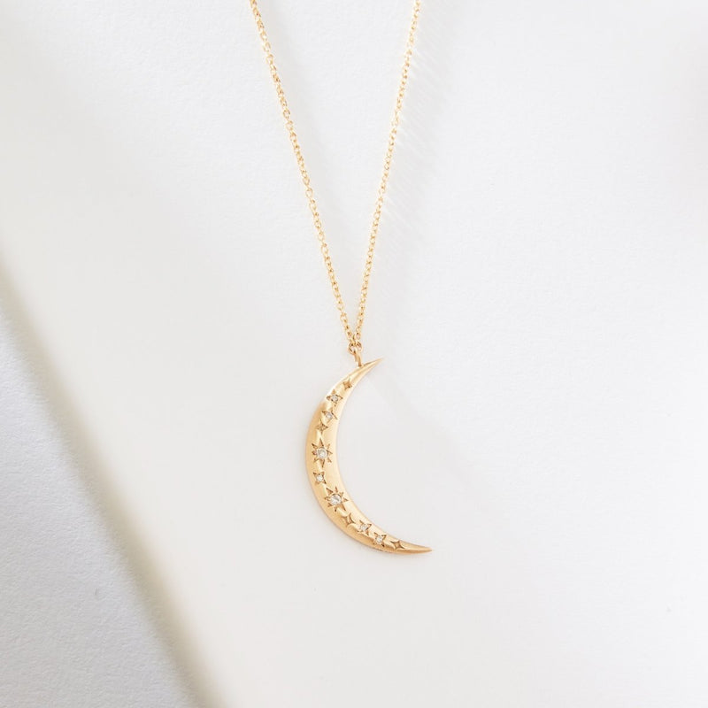 A delicate crescent moon tie dye necklace crafted from 14k yellow gold by bluboho features a pendant adorned with small star engravings. Displayed against a clean, white background, the necklace highlights its elegant and charming design.