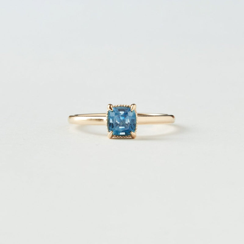 The daydream bliss one-of-a-kind ring by beloved by bluboho, featuring a cushion-cut blue sapphire set in 14k yellow gold, is displayed against a plain white background. The simple, elegant design highlights the vibrant blue stone.