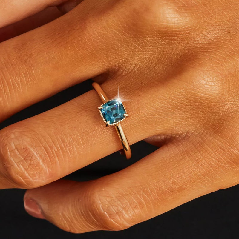 A close-up photo showcases a hand adorned with the "daydream bliss one-of-a-kind ring" by beloved by bluboho, featuring 14k yellow gold and a cushion cut blue sapphire. The ring is worn on the ring finger, with the gemstone catching the light to create a subtle sparkle. The dark background accentuates both the hand and the exquisite ring.