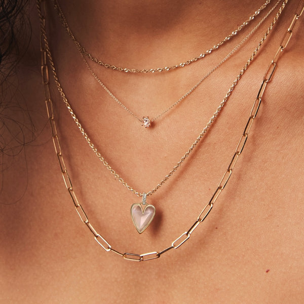 A close-up of a person's neck wearing three layered gold necklaces. The shortest necklace features a small gemstone, the middle one showcases bluboho's pink opal diamond heart mood charm made with 10k yellow gold, pink opal, and diamonds, and the longest one has elongated chain links.