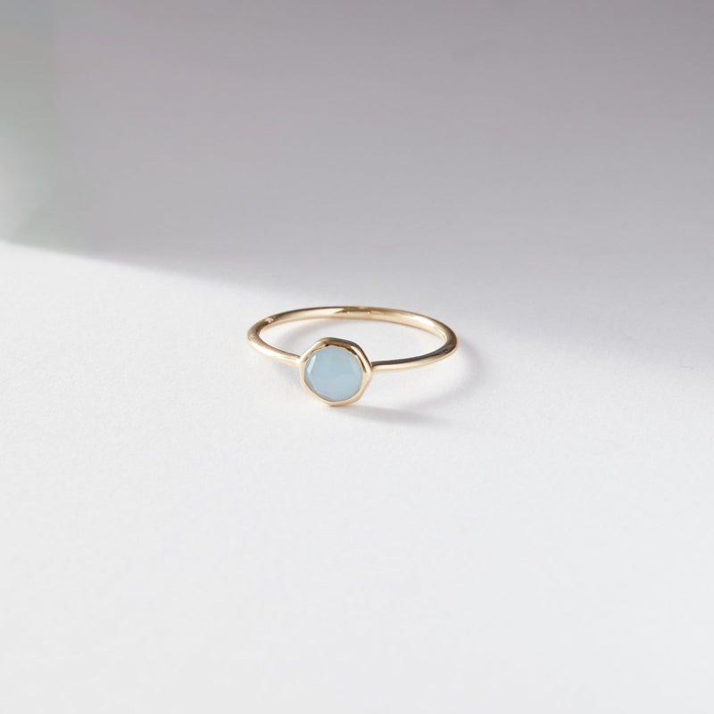 The aquamarine mini mood ring from bluboho, crafted in 10k yellow gold and featuring a round pale blue aquamarine gemstone, is displayed on a white surface. The ring casts a soft shadow diagonally across the background, highlighting its minimalistic and elegant design.