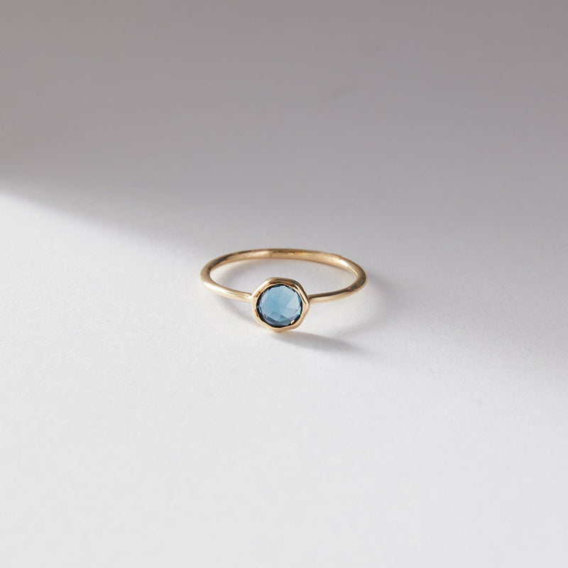 The london blue topaz mini mood ring by bluboho, crafted in 10k yellow gold and featuring a single hexagonal blue topaz gemstone, is showcased on a light gray surface with a soft shadow cast to the left, portraying a minimalist and elegant jewelry design.