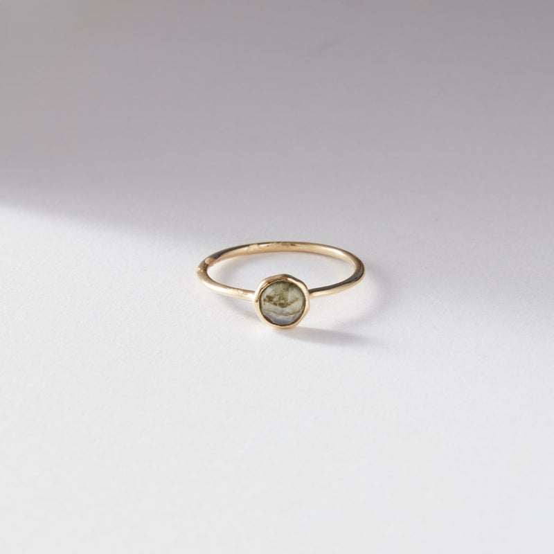 A 10k yellow gold labradorite mini mood ring from bluboho lies on a white surface, illuminated by soft, diffused light. The ring showcases a simple round labradorite stone set in the center. Shadows and light create a subtle gradient in the background.