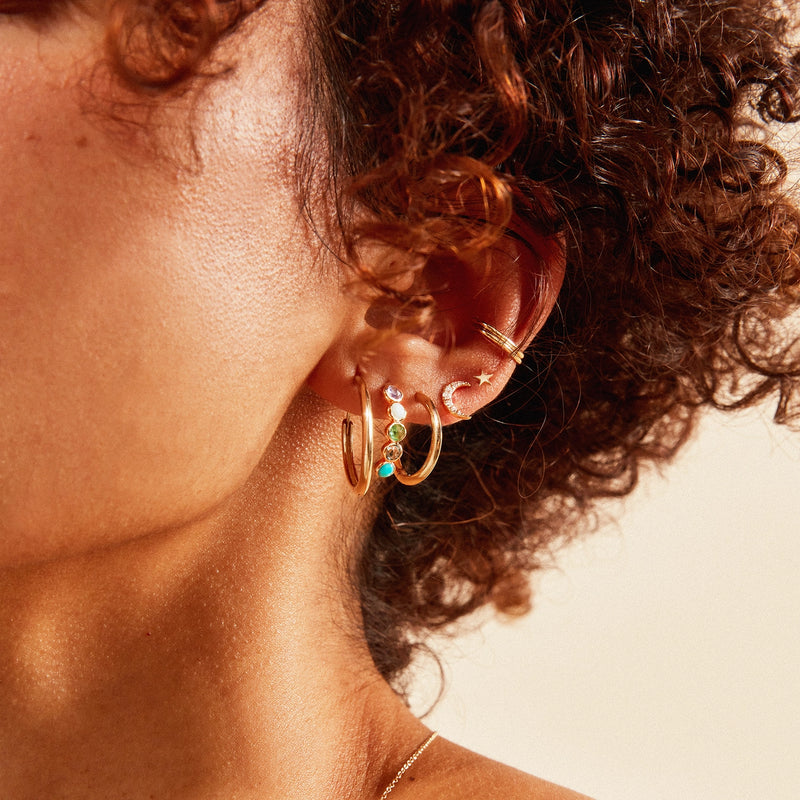 Close-up of a person's ear adorned with multiple gold earrings. There are three hoop earrings, one of which is the bluboho coastal nomad five stone mood hoop set in 14k yellow gold and featuring tanzanite, larimar, emerald, London blue topaz, and turquoise stones. The other two hoops feature turquoise beads. Additionally, there's an earring shaped like a crescent moon. The person has curly hair and a subtle complexion.
