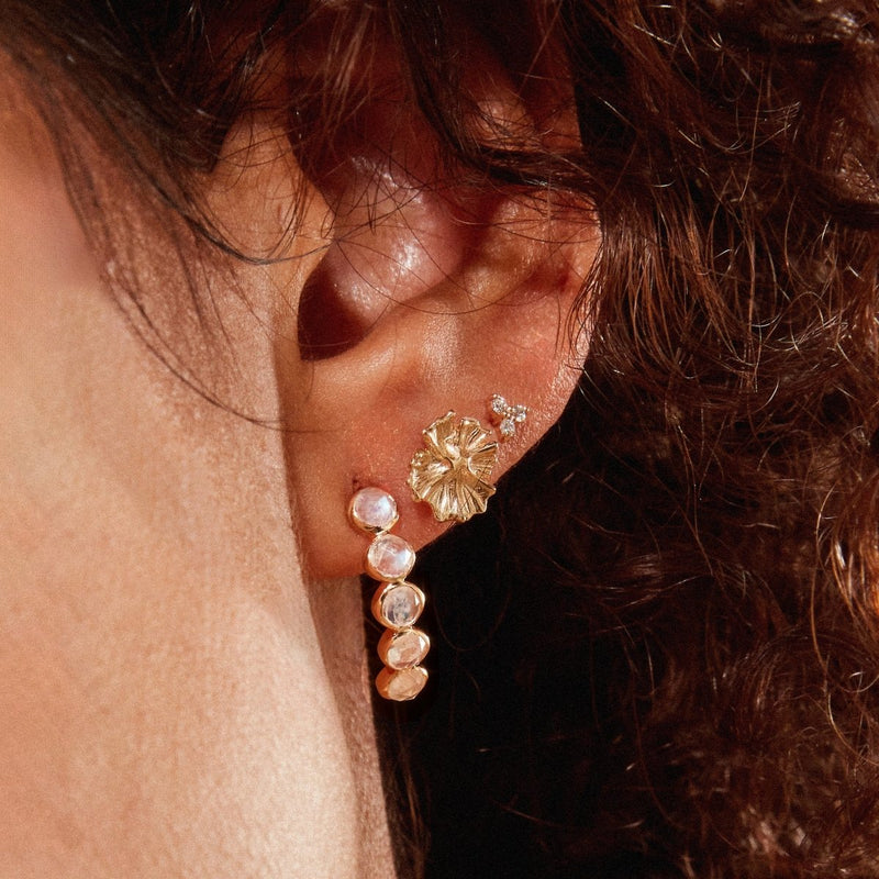 Close-up of an ear adorned with a bluboho five stone moonstone mood hoop in 14k yellow gold, with the hoop design extending along the curve of the ear and featuring several moonstones that resemble pearls. Higher up, there's a single small stud diamond earring. The background shows curly hair.