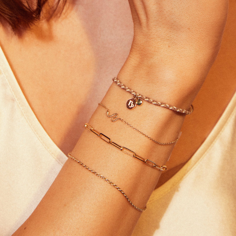 A close-up of a person's wrist adorned with five delicate bracelets in various styles, including a braided cord, one with a small charm featuring a "B", a thin gold chain, a bracelet with a snake design, and the capricorn zodiac earth element cord bracelet - 10k yellow gold, emerald, cord by bluboho. The person is wearing a sleeveless top.