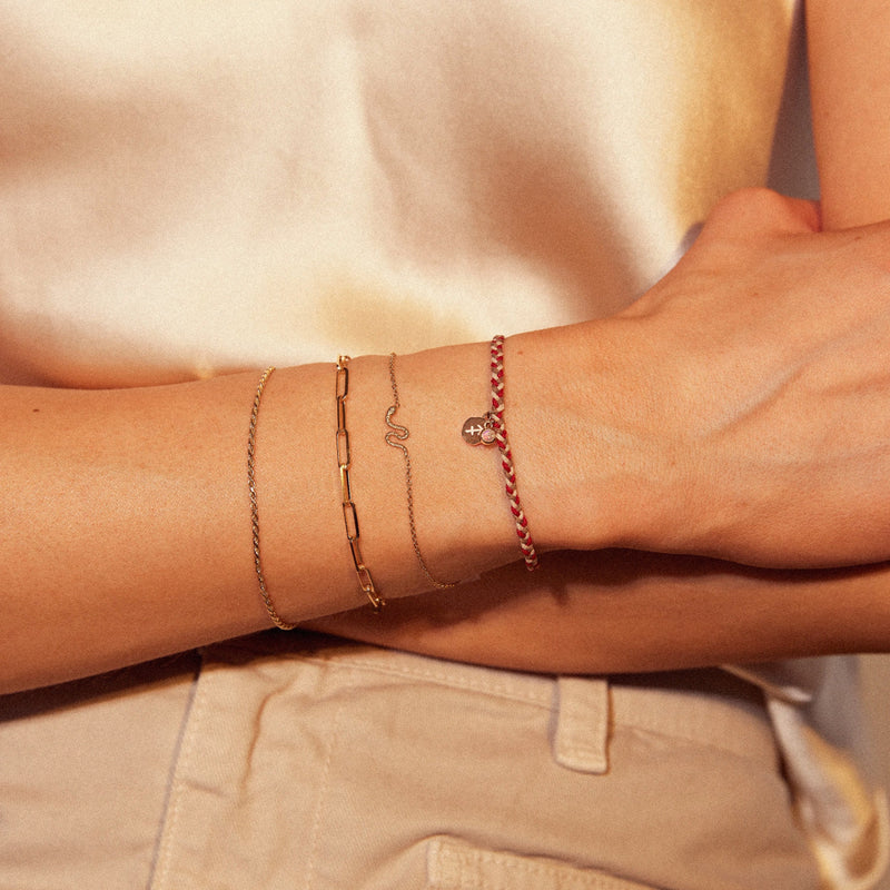 A close-up of a person's left wrist adorned with four different bracelets, including the leo zodiac bracelet fire element cord bracelet - 10k yellow gold, opal, cord from bluboho. The bracelets also include a thin chain, a snake-shaped chain, a geometric rectangular chain, and a red braided chain with a small charm. The person is dressed in a light-colored top and pants.