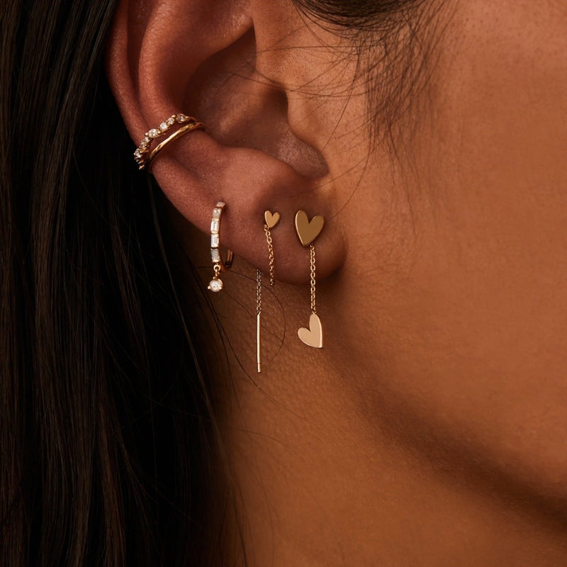 Close-up of a person's ear adorned with multiple earrings. The upper ear features a gold hoop with small jewels. The lower earlobe has three earrings: a small heart stud, a dangle earring with a heart charm, and **bluboho's lovely heart threader earring - 14k yellow gold**.