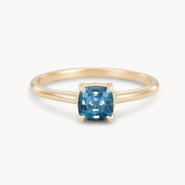 The Daydream Bliss One-of-a-Kind Ring by Beloved by Bluboho showcases a 14k yellow gold band adorned with a blue cushion-cut sapphire held securely by four prongs. The faceted surface of the gemstone enhances its sparkle and elegance, set against a plain white background.