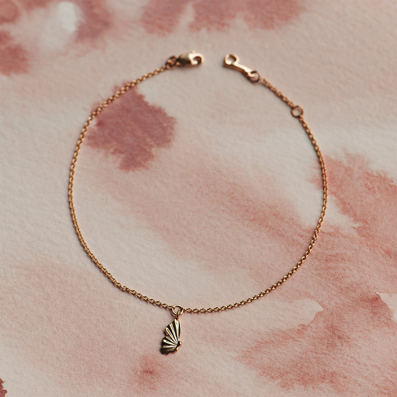 The bluboho metamorphosis butterfly wing bracelet, crafted from 14k yellow gold and fastened with a fine chain and lobster clasp, is elegantly displayed against a pink and beige watercolor background.