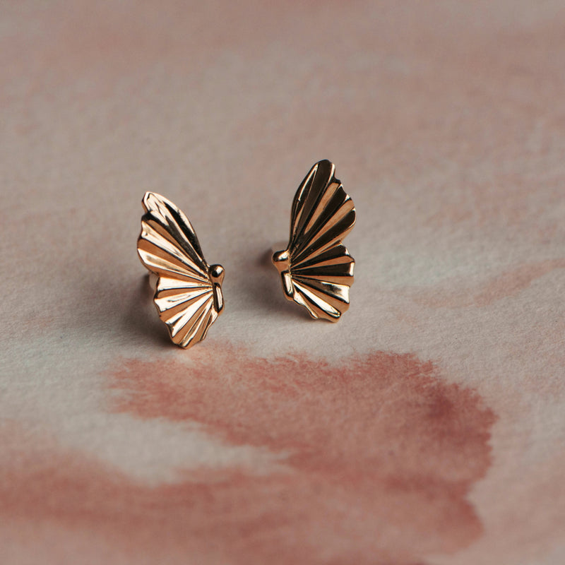 The metamorphosis butterfly wing earrings from bluboho, crafted in 14k yellow gold, are showcased on a soft, pinkish background. The delicate design of these earrings features intricate lines that mimic the natural form of butterfly wings.