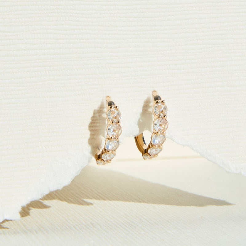 A pair of bluboho's Large Moonglade hoop earrings with embedded round, clear gemstones is displayed against a textured, off-white background. The earrings are secured around the top edge of a piece of torn white paper, showcasing their intricate detailing and elegant design.