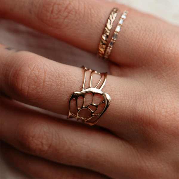 Close-up of a hand adorned with three gold rings. The middle finger features the distinctive winged being butterfly ring by bluboho, crafted from 14k yellow gold in an open-work design that resembles abstract lines and shapes. Simpler, narrow band rings with textured details grace the index and pinky fingers.