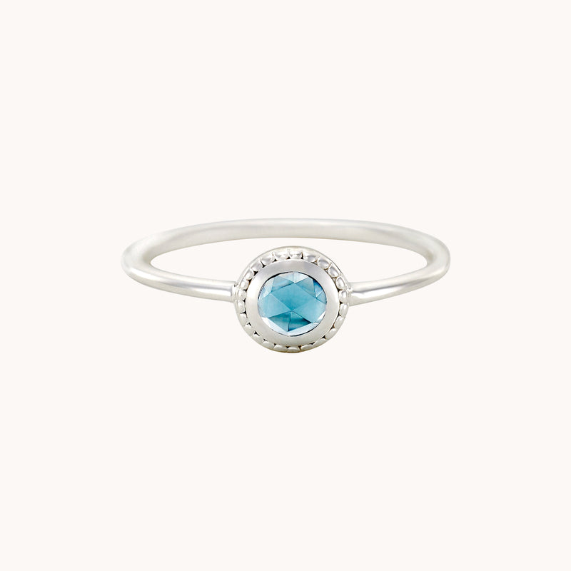 Introducing the full moon London blue topaz ring from bluboho, crafted in sterling silver. This delicate ring showcases a striking round, faceted blue topaz gemstone at its center, encased within a textured, circular bezel setting. The design features a simple, thin band that embodies elegance and minimalism.