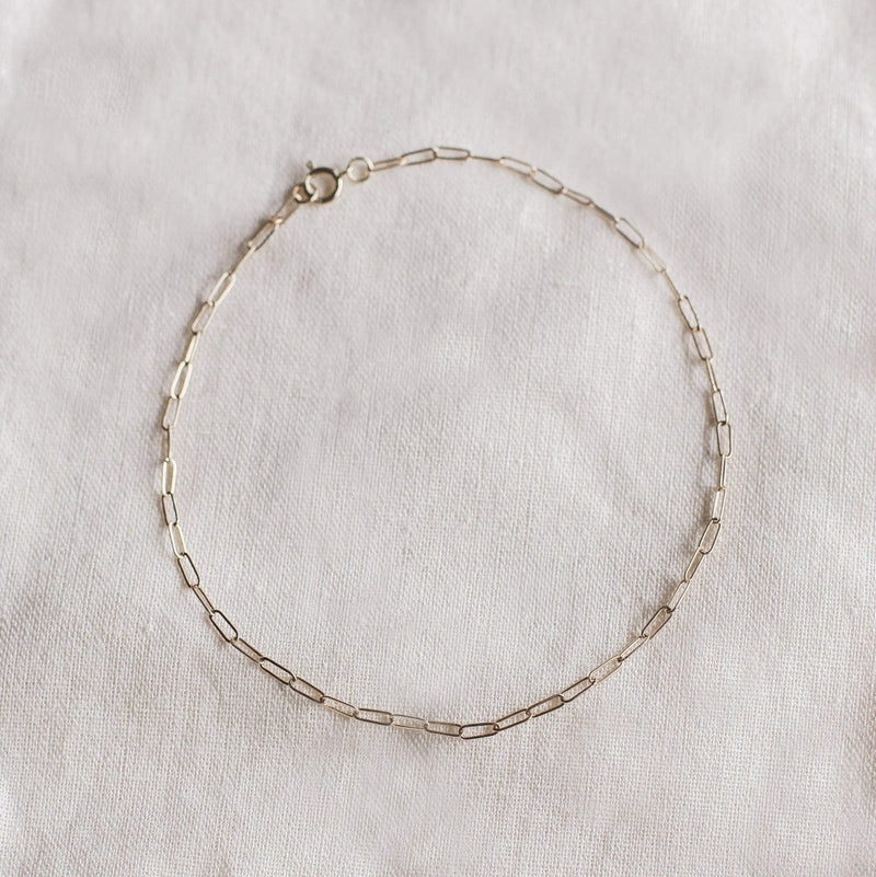 The Infinite Inseparable Anklet by bluboho, crafted in 14k yellow gold, features a delicate elongated link design. It is displayed on a light, textured fabric background and is secured with a small clasp.