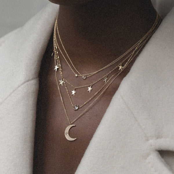 A person wears multiple gold necklaces layered elegantly, including the Moonlight Crescent Diamond Necklace by bluboho. The necklaces feature various charms, such as a crescent moon, stars, and small gemstones, all resting against a beige coat. The neutral background highlights the delicate jewelry beautifully.