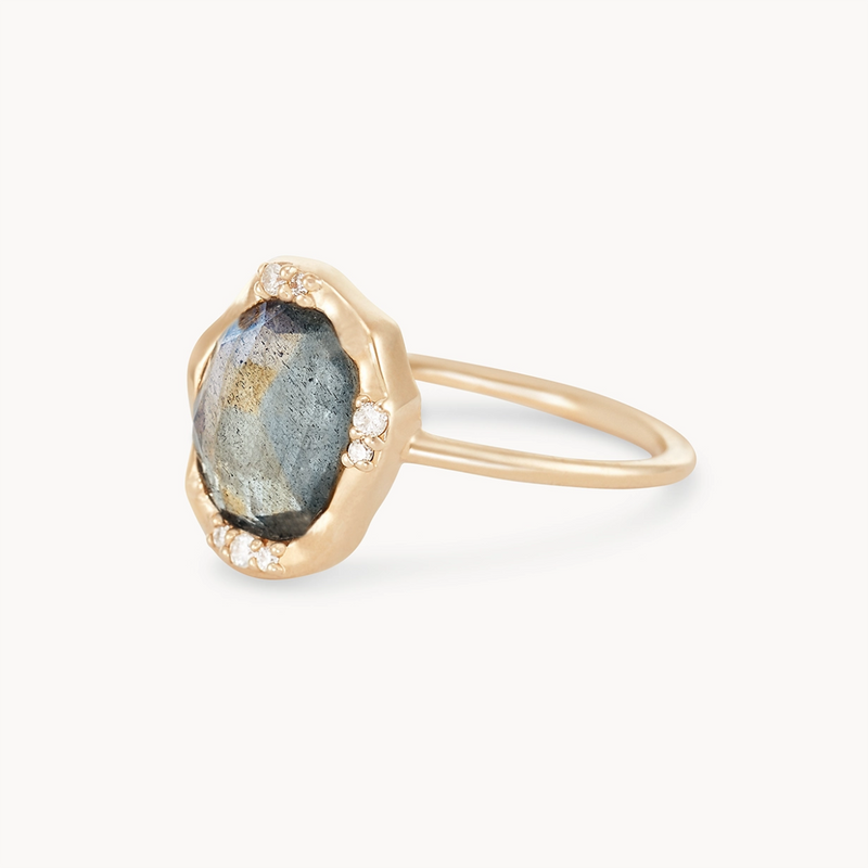 A close-up image of bluboho's elegant labradorite mood ring, crafted in 14k yellow gold, featuring a large, faceted dark labradorite gemstone at its center. The centerpiece is surrounded by small, sparkling white stones set in a wavy gold band. The thin band showcases the intricate design of the centerpiece beautifully.