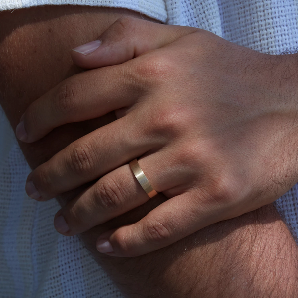 A close-up of a person's hands crossed at the wrist, adorned with the "Larger Everlast Band Brushed" by beloved by bluboho—a simple, brushed 14k yellow gold band gleaming on the ring finger of their left hand. The person is dressed in a white textured fabric, possibly a shirt or sweater. The lighting enhances the intricate details of both their hands and the elegantly crafted ring.