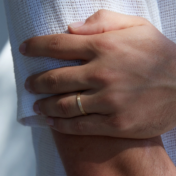 Close-up of a person's hands where they are wearing the Everlast band polished—14k yellow gold, polished—by beloved by bluboho on the ring finger of the left hand while their right hand rests on their left arm. The person is dressed in a textured white garment with sunlight casting gentle shadows.