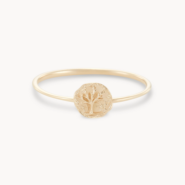 The "tiny tree of life ring - 14k yellow gold" by bluboho is a delicate gold ring adorned with a small round disc at its center, intricately engraved with a tree design. This minimalist and elegant piece is showcased against a plain white background.