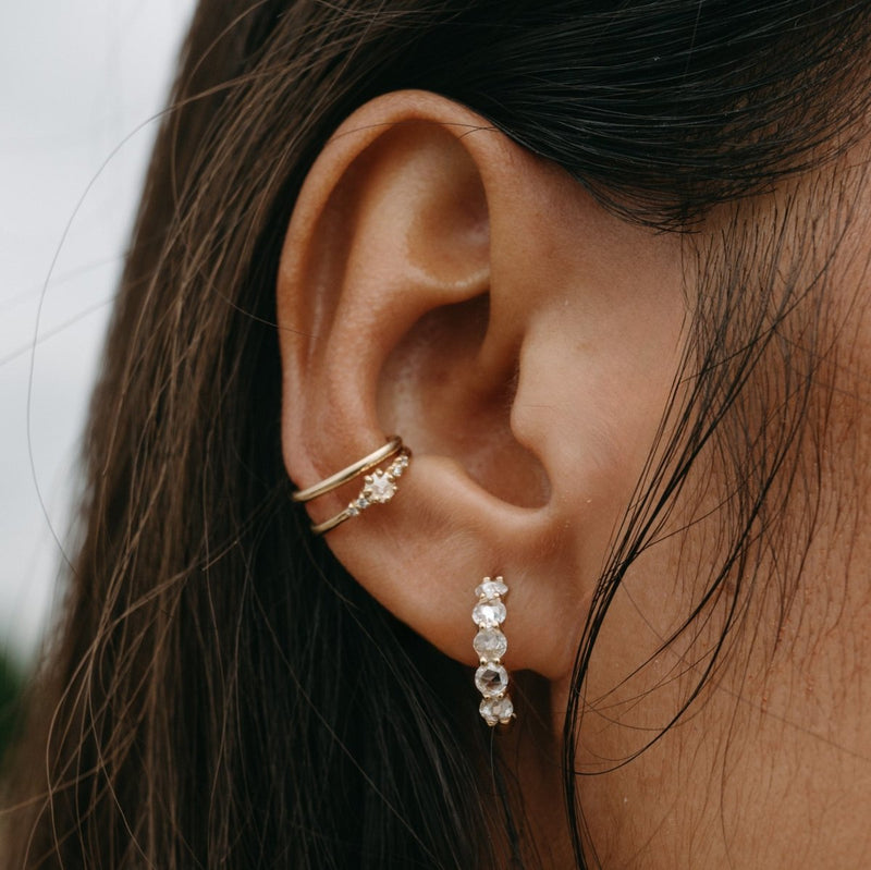 Close-up of a woman's ear adorned with two types of earrings. One is the "nova ear cuff" in 14k yellow gold by bluboho, featuring a floral design on the upper part of the ear (helix), and the other is a dangling earring with multiple clear gemstones on the earlobe. The woman's hair is partially covering her ear.