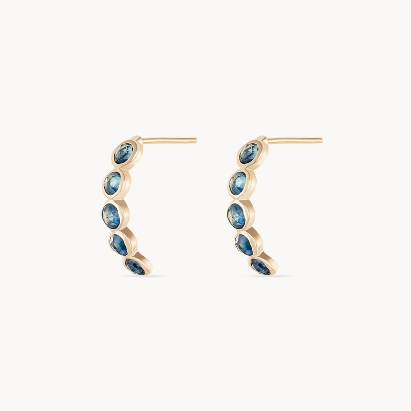 The image features a pair of bluboho blue sapphire five stone mood hoops in 14k yellow gold. The earrings are adorned with multiple blue sapphires and are positioned facing forward, beautifully showcasing their semi-circular design and the arrangement of the stones along the hoops. The background is plain white.