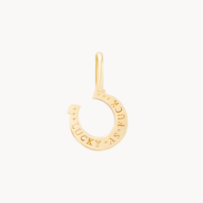 The wild luck horseshoe charm by bluboho is a small, 10k yellow gold pendant featuring the phrase "LUCKY AS FUCK" engraved on it. The charm has a loop at the top for attaching to a chain and is showcased against a plain white background.