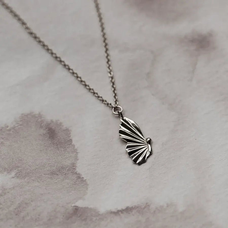 A delicate silver chain necklace featuring a butterfly pendant with intricately designed wings. The metamorphosis butterfly wing necklace - sterling silver by bluboho is laid out flat on a soft, marble-patterned background.