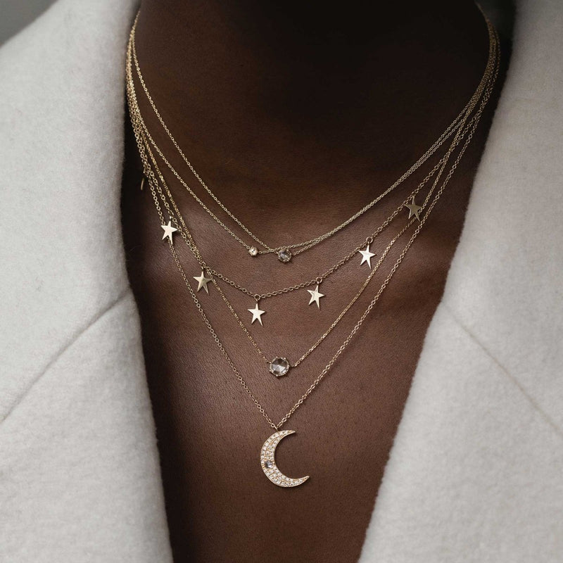 A close-up of a person wearing a light-colored, soft-textured garment. They have layered necklaces, including the large supernova necklace by bluboho, featuring a crescent moon pendant, small star charms, and a few delicate chains with small jewels.