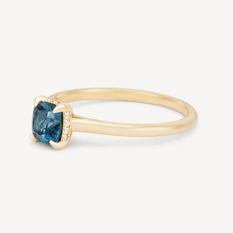 The daydream bliss one-of-a-kind ring by beloved by bluboho showcases a large, cushion-cut blue sapphire in a prong setting, accented with small diamonds on either side of the central stone. The 14k yellow gold band is thin and smooth, accentuating the elegant design of the ring.