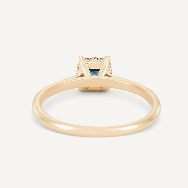 A gold ring called the "daydream bliss one-of-a-kind ring" by beloved by bluboho features a solitaire cushion cut blue sapphire set in a four-prong setting on a simple 14k yellow gold band. The elegantly designed ring prominently showcases the gemstone against a plain white background, highlighting its exquisite color and design.