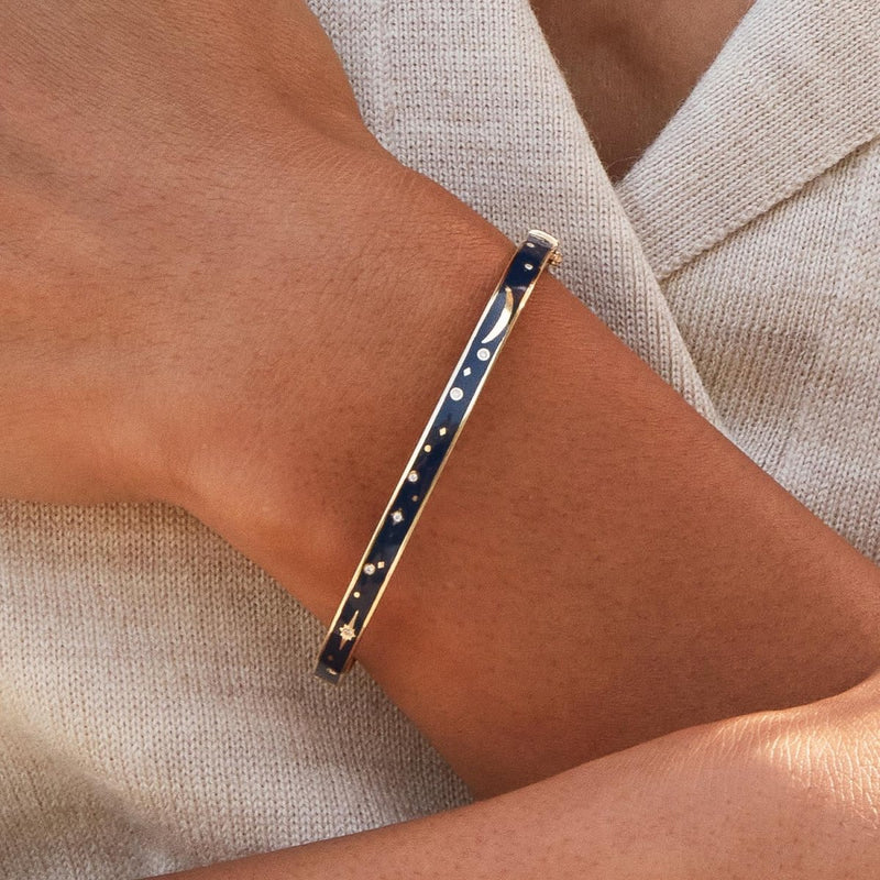 A close-up of a person's wrist adorned with bluboho's "the dream state bangle," a slim bracelet made of 14k yellow gold with a dark blue enamel design featuring small star and moon shapes and dots. The person is dressed in light beige, textured fabric.