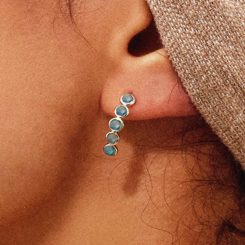 Close-up of an ear adorned with a blue sapphire five stone mood hoop from bluboho, featuring a vertical line of five small, round blue sapphires set in a 14k yellow gold frame. The person is wearing a beige knitted cap that partially covers the ear.