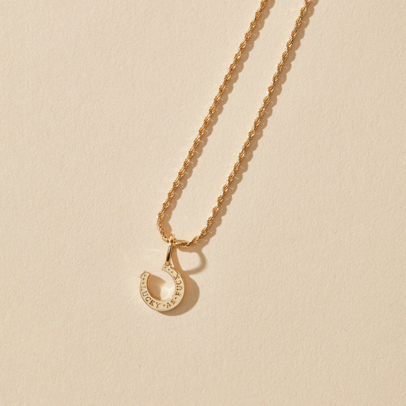 A 10k yellow gold, diamond wild luck horseshoe charm from bluboho lies on a beige background. The pendant features an intricate design resembling a hollow crescent moon, engraved with the text "MULBERRY" and "AR RUD," separated by a small star.