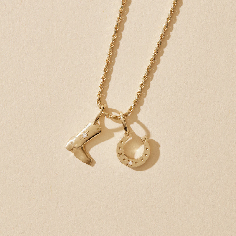 A detailed 10k yellow gold necklace from bluboho, featuring two charms: a cowboy boot adorned with tiny diamond star embellishments and a horseshoe, elegantly displayed against a neutral background.