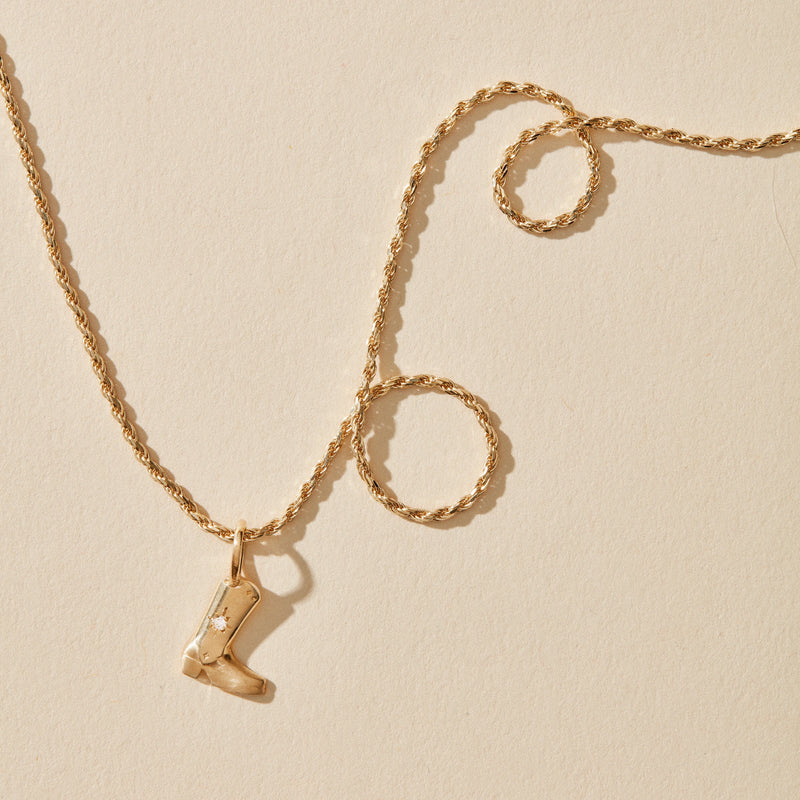 An abundance boot charm necklace from bluboho, crafted in 10k yellow gold and adorned with a delicate twisted chain, is artfully displayed on a light beige background. The pendant is an intricately detailed cowboy boot featuring a tiny embedded diamond near the top. The chain forms gentle loops beside the pendant.