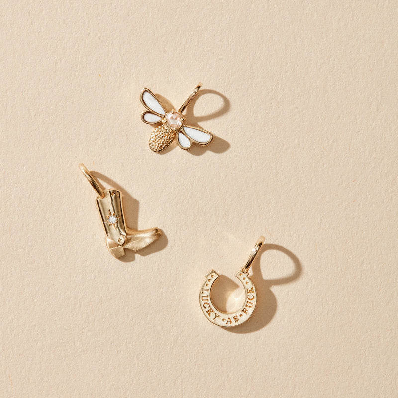 A trio of gold pendants on a beige background: the honey glow bee charm featuring 10k yellow gold with a mother of pearl inlay from bluboho, a cowboy boot with a star, and a horseshoe inscribed with "Luck." Each charm is finely detailed, evoking themes of nature, western style, and good fortune.