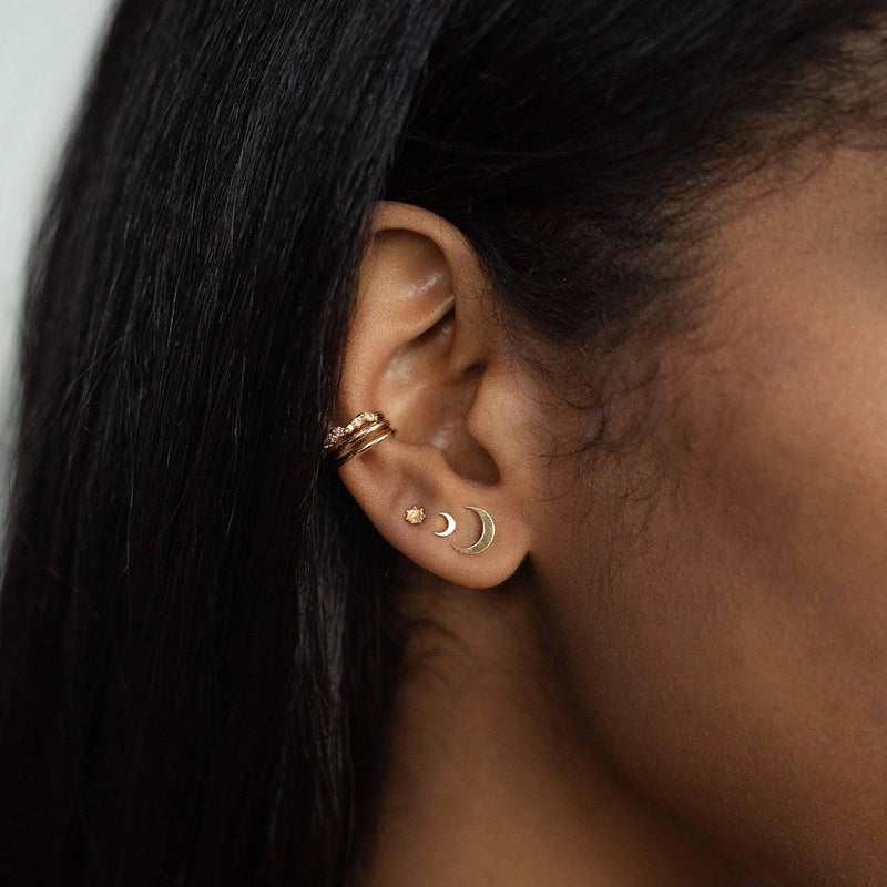 A close-up photo of a person’s ear adorned with multiple gold earrings, including the bluboho "Everyday larger crescent moon earring" in 14k yellow gold, a small star, and two delicate bands on the upper part of the ear. The person’s straight black hair is partially visible.