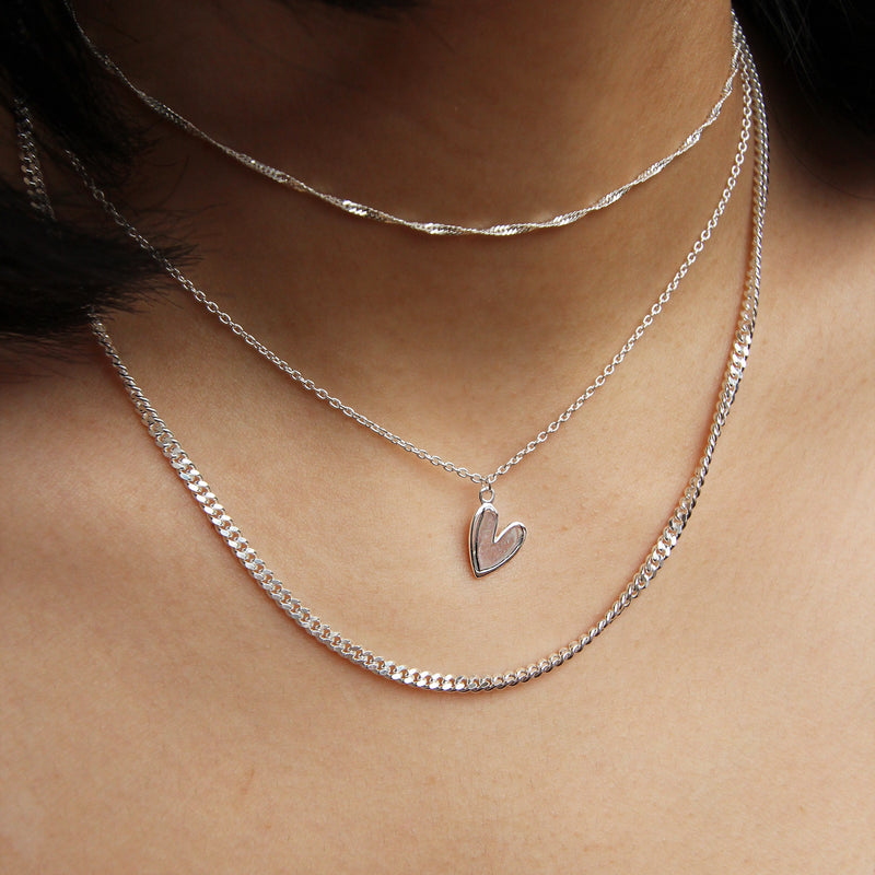 Close-up of a person wearing three silver necklaces. The shortest necklace is a thin chain, the medium one features a small, hollow heart pendant, and the longest one is the "curb silver necklace - sterling silver" by bluboho, with its flat, wide link chain draping below the collarbone. The background is blurred skin.