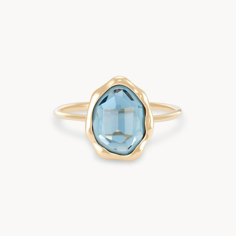 A london blue topaz mood ring by bluboho, crafted from 14k yellow gold, showcases an oval-cut blue topaz gemstone secured by a wavy, organic bezel setting. The faceted gemstone sits atop a simple and delicate band. The ring is set against a plain white background.