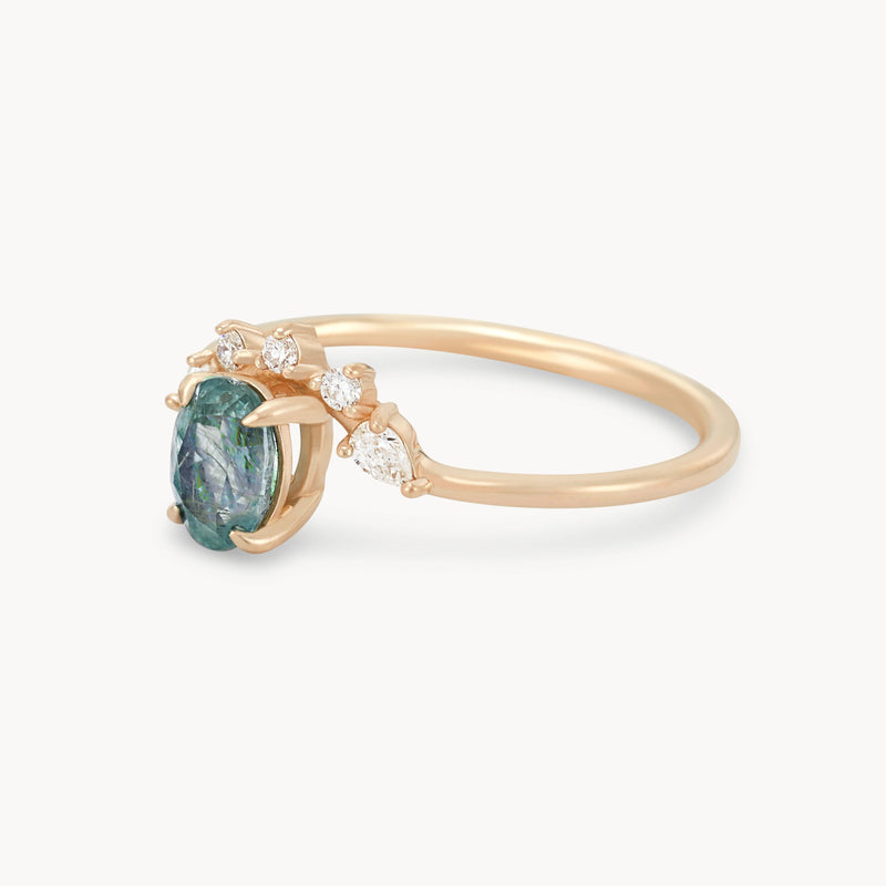 The Love’s Unity One-Of-A-Kind ring by Beloved by Bluboho is a delicate 14k yellow gold piece that features a cyan green oval sapphire gemstone at its center. It's elegantly complemented by three small white diamonds positioned on one side, with the polished gold band adding an elegant touch to the design.
