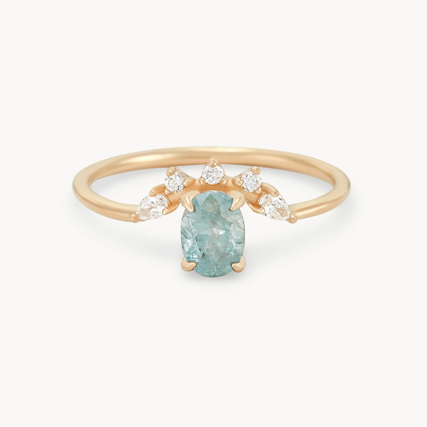 Introducing the Love’s Unity One-of-a-Kind ring by Beloved by Bluboho: a striking piece crafted from 14k yellow gold, featuring a central cyan green oval sapphire. Flanked by three small teardrop-shaped diamonds on either side, the thin and simple band elegantly highlights the gemstones against a plain white background.