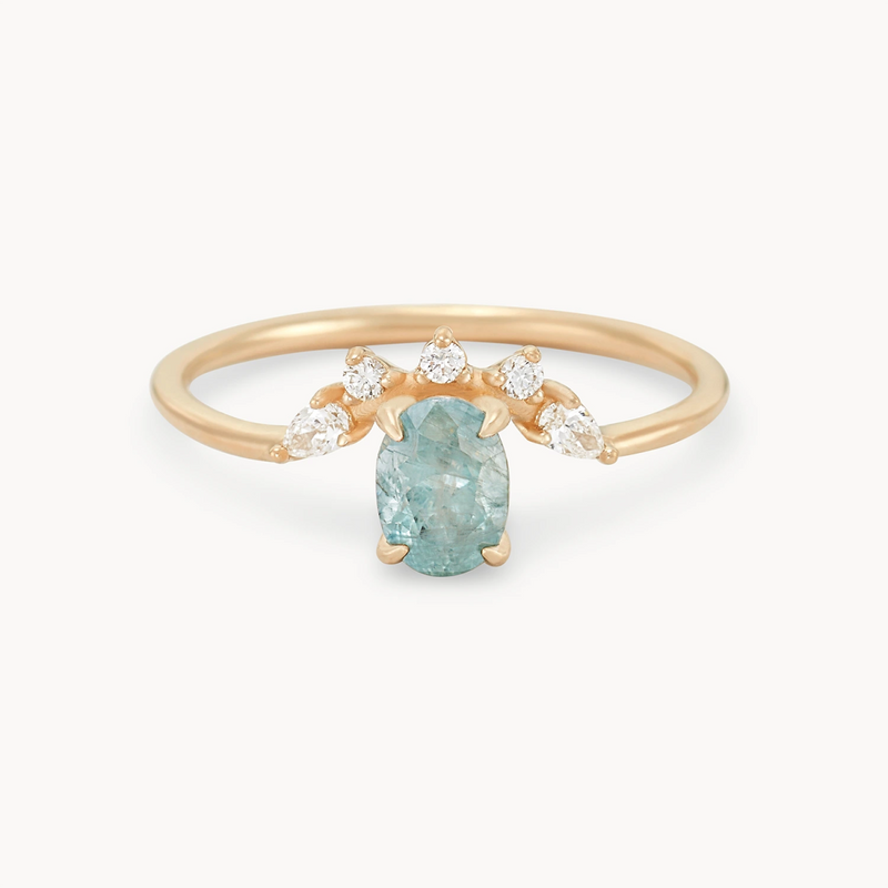 Introducing the Love’s Unity One-of-a-Kind ring by Beloved by Bluboho: a striking piece crafted from 14k yellow gold, featuring a central cyan green oval sapphire. Flanked by three small teardrop-shaped diamonds on either side, the thin and simple band elegantly highlights the gemstones against a plain white background.