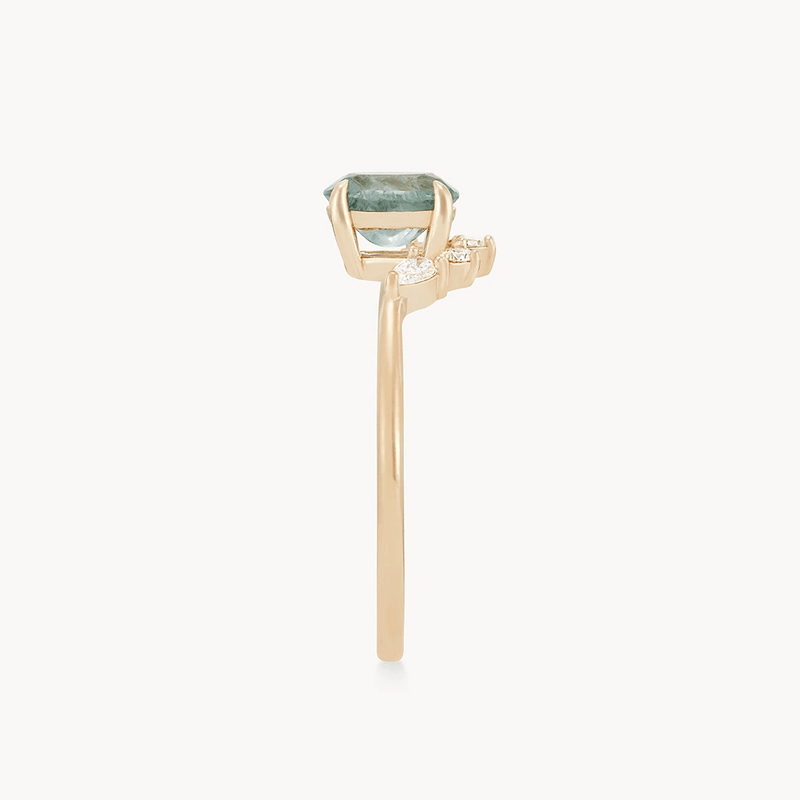 A close-up side view of the Love’s Unity one-of-a-kind ring by beloved by bluboho, showcasing a 14k yellow gold band with a cyan green oval sapphire set in a prong setting. The slim and polished band is accented with two small round diamonds on either side of the main stone. The background is plain and white.