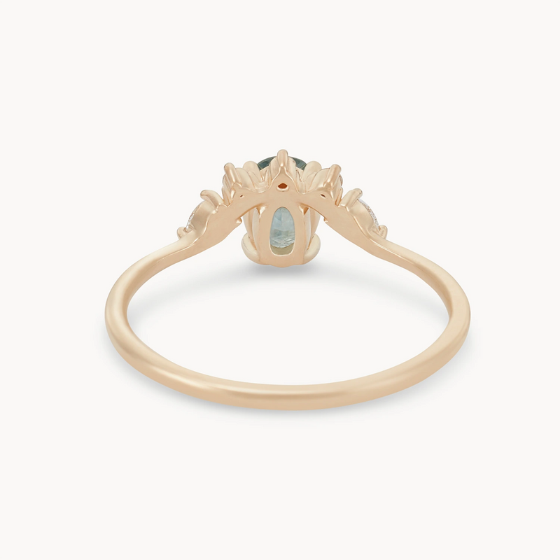 The Love’s Unity One-of-a-Kind ring by beloved by bluboho is crafted from 14k yellow gold and showcases an exquisite cyan green oval sapphire at its center. The side view reveals intricate detailing on the band, flanked by smaller stones, and the design features open spaces beneath the prominent gemstone setting to highlight its beauty.