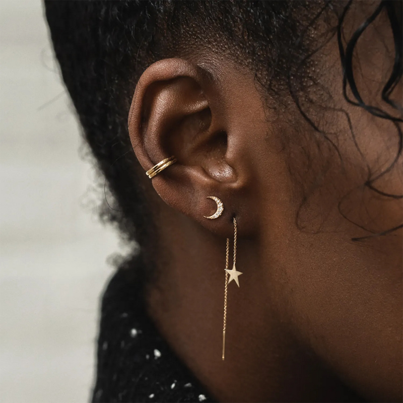 A close-up of a person's ear adorned with two bluboho gold earrings: a Moonlight crescent earring in 14k yellow gold and a dangling star earring attached to a thin chain. The person's ear also features a small gold cuff. The background is out of focus.