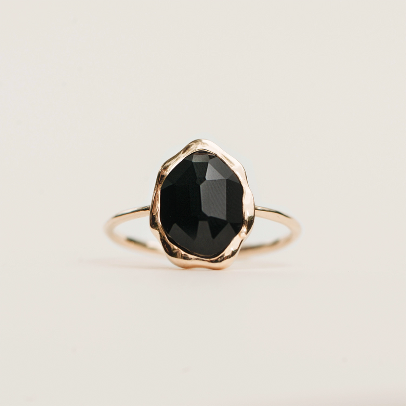 A delicate black onyx mood ring - 14k yellow gold from bluboho, featuring a large faceted round black gemstone at its center. The polished gemstone is beautifully surrounded by a thin gold band that enhances its elegance, set against a neutral light beige background.